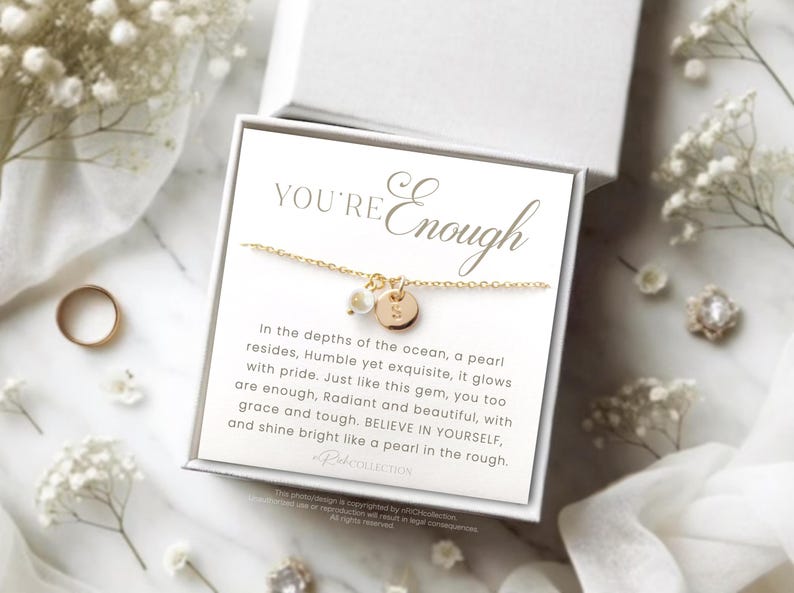 May include: A gold necklace with a pearl and a small gold disc charm. The necklace is displayed on a white card with the text "YOU'RE Enough" and a quote about pearls.