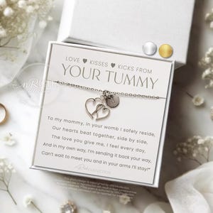 Op de afbeelding: Een zilveren armband met twee hartbedels en een kleine ronde bedel, gepresenteerd op een witte kaart met de tekst "LOVE KISSES KICKS FROM YOUR TUMMY". De kaart zit in een witte doos, omringd door witte bloemen en een gouden ring.