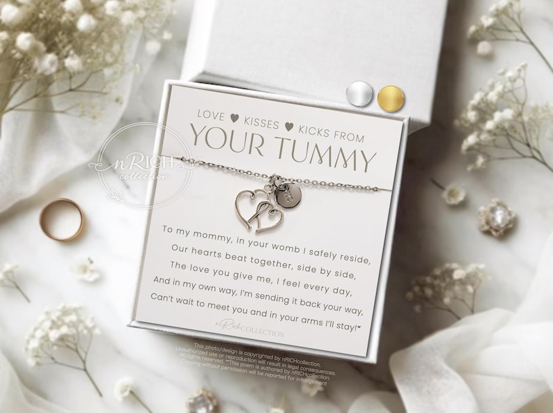 Op de afbeelding: Een zilveren armband met twee hartbedels en een kleine ronde bedel, gepresenteerd op een witte kaart met de tekst "LOVE KISSES KICKS FROM YOUR TUMMY". De kaart zit in een witte doos, omringd door witte bloemen en een gouden ring.