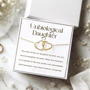 May include: A white card with the text "Unbiological Daughter" and a message about a necklace with two intertwined hearts. The message reads, "You may not be my daughter by birth, but you are my daughter by heart. Wear this necklace as a symbol of the love, pride, and unbreakable bond we share - two hearts forever connected."