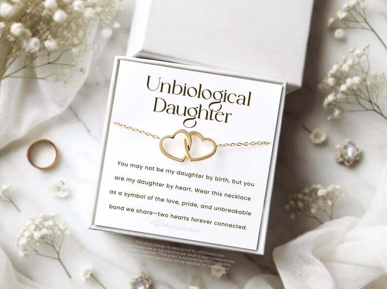 May include: A white card with the text "Unbiological Daughter" and a message about a necklace with two intertwined hearts. The message reads, "You may not be my daughter by birth, but you are my daughter by heart. Wear this necklace as a symbol of the love, pride, and unbreakable bond we share - two hearts forever connected."