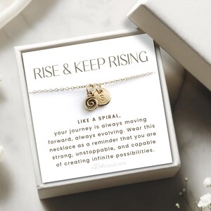 May include: A gold necklace with a spiral charm and a heart charm. The necklace is in a white box with a card that reads "Rise & Keep Rising" and a message about the journey being like a spiral.