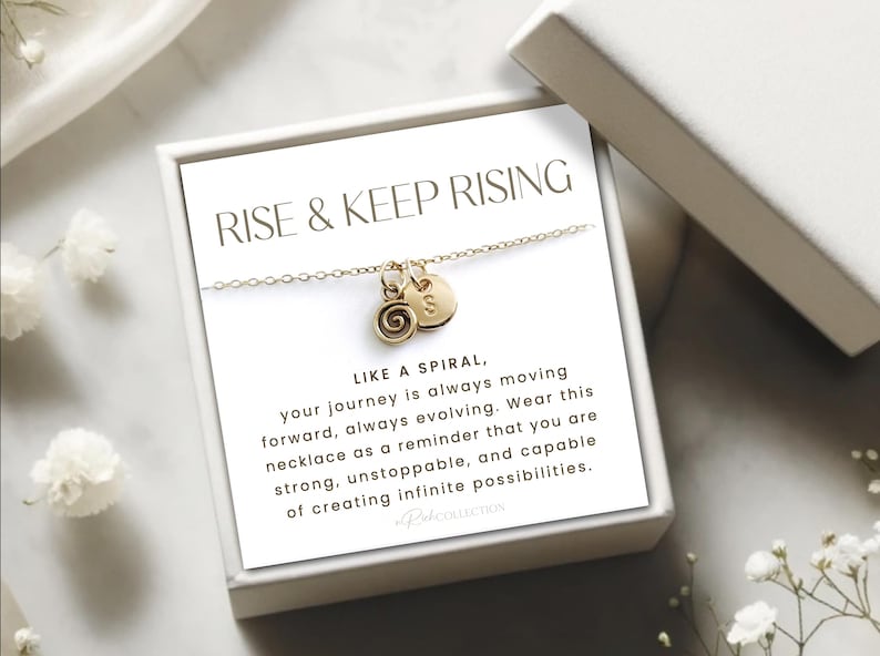 May include: A gold necklace with a spiral charm and a heart charm. The necklace is in a white box with a card that reads "Rise & Keep Rising" and a message about the journey being like a spiral.