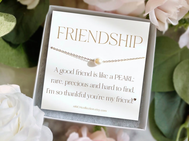 Może przedstawiać: Biała kartka z pozłacanymi napisami zawiera tekst "FRIENDSHIP" i "A good friend is like a PEARL: rare, precious and hard to find. I'm so thankful you're my friend!" Złoty łańcuszek z pojedynczą perłą zwisa nad tekstem. Kartka znajduje się w białym pudełku.