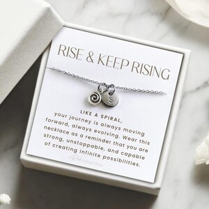 May include: A silver necklace with a spiral charm and a letter charm in a white box. The box has the text "RISE & KEEP RISING" and a message about the necklace being a reminder to be strong and capable of creating infinite possibilities.