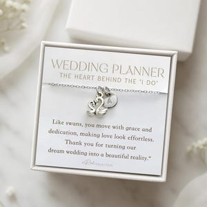 May include: A silver necklace with a swan charm and an initial pendant, presented in a white gift box. The box contains a card with text that reads "WEDDING PLANNER" and a thank you message. The necklace is a wedding gift.