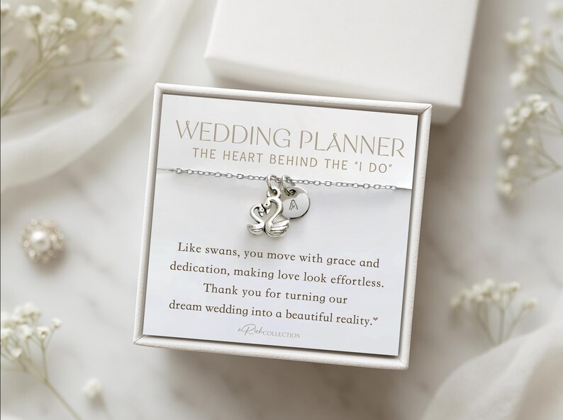 May include: A silver necklace with a swan charm and an initial pendant, presented in a white gift box. The box contains a card with text that reads "WEDDING PLANNER" and a thank you message. The necklace is a wedding gift.