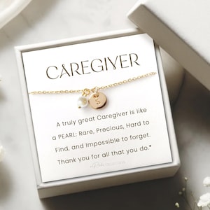 May include: A white box with a gold necklace inside. The necklace has a pearl charm and a gold disc charm with the letter "S" on it. The box has a message that reads "A truly great Caregiver is like a PEARL: Rare, Precious, Hard to Find, and Impossible to forget. Thank you for all that you do."