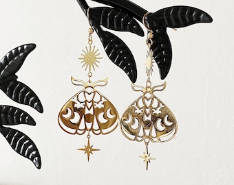 Può includere: Orecchini dorati con un designo di falena dettagliato con accenti di stella e mezzaluna. Gli orecchini sono appesi a un espositore per gioielli nero.