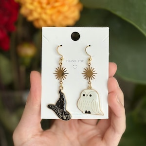 Puede incluir: Un par de pendientes colgantes dorados con un dije de sol, uno con un sombrero de mago negro con el texto WIZARD IN TRAINING y el otro con un búho blanco. Los pendientes se presentan en una tarjeta blanca con un orificio en la parte superior.