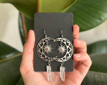 May include: A pair of silver-toned dangle earrings featuring a crescent moon cutout design with a small sun charm suspended in the center. Each earring is finished with a clear, raw crystal point hanging from the bottom of the moon shape.