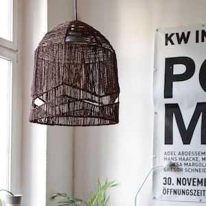 Könnte beinhalten: Eine braune, gewebte Pendelleuchte mit konischem Schirm. Der Schirm hat ein geometrisches Design mit offenen Abschnitten. Das Licht hängt an einem schwarzen Kabel. Der Hintergrund umfasst ein Fenster und ein Poster mit Text.