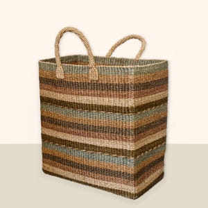 Puede incluir: Una cesta de almacenamiento grande, rectangular y tejida con dos asas. La cesta está hecha de fibras naturales y tiene un patrón de rayas en tonos marrones, beige y verdes.
