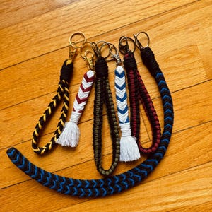 May include: A selection of handmade keychains with braided cords in various colours, including black, yellow, red, white, blue, and green. Each keychain features a metal clasp and a decorative tassel. The keychains are arranged on a wooden surface.