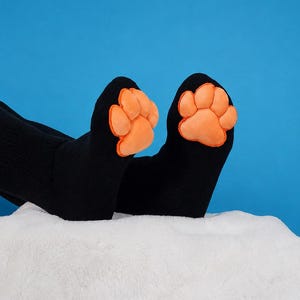 Könnte beinhalten: Ein Paar schwarze Socken mit weichen, orangefarbenen, gepolsterten Pfotenabdrücken an den Zehen. Die Socken bestehen aus einem weichen, einfarbig schwarzen Stoff und reichen bis über den Knöchel, wobei die leuchtend orangefarbenen Zehenpolster hervorstechen.