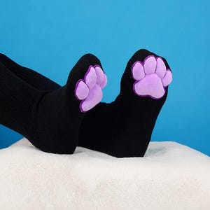 May include: A pair of black socks featuring a plush, light purple paw print design on the toe area. The socks are made of a solid black knit fabric, and the paw pads are stitched onto the front of each sock, creating a raised, textured appearance.