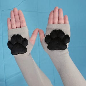 May include: A pair of light grey fingerless gloves featuring a black paw print on the palm. The gloves are made from a soft, stretchy material, designed to cover the hand and wrist.