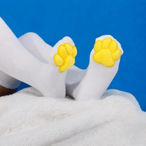 May include: A pair of white socks featuring bright yellow, padded paw-shaped appliques on the toe area. The socks are made of a smooth, solid white fabric and the paw designs consist of a large central pad with four smaller rounded toe pads.