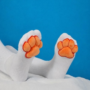 May include: A pair of white socks featuring plush, orange, padded paw-shaped appliqués on the front. The paw designs have distinct toe pads and a central pad outlined in red stitching, creating a textured, three-dimensional appearance on the fabric.