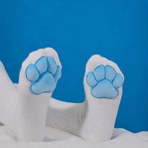 May include: A pair of white socks featuring a plush, light blue animal paw print design on the sole. The paw print consists of a large central pad and four smaller toe pads, all rendered in a soft, textured fabric against the white knit material.