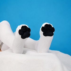 May include: A pair of white knee-high socks featuring black, plush, padded paw-shaped appliques on the front of the feet. The socks are made of a knit fabric and are positioned to display the black paw pads clearly against a solid blue background.
