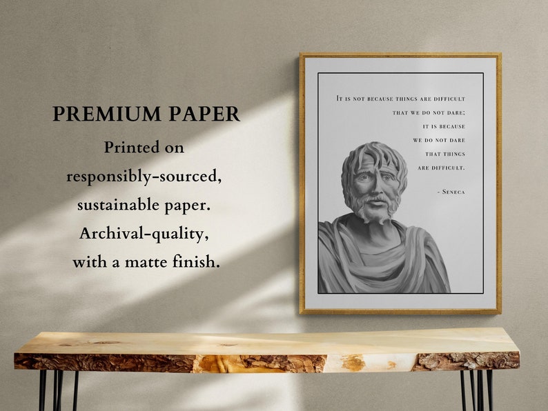 May include: A framed print with a black and white image of a man's bust. The print has a quote by Seneca: "It is not because things are difficult that we do not dare; it is because we do not dare that things are difficult." The print is on a wooden table with a light wood grain.