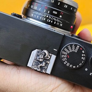 May include: A black rangefinder camera with a silver lens and a detailed dial. The camera has a silver decorative plate with an ornate design. The lens has various settings and measurements. The camera is held in a hand.