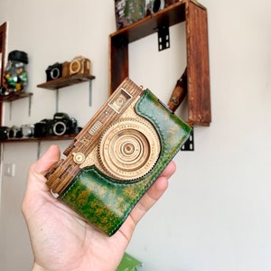 Puede incluir: Una cámara de madera y cuero con un exterior de cuero verde y una lente de madera. La cámara tiene una lente circular con el texto "SUPER EBC FUJINON 1:2.7 28mm" impreso alrededor de la lente.