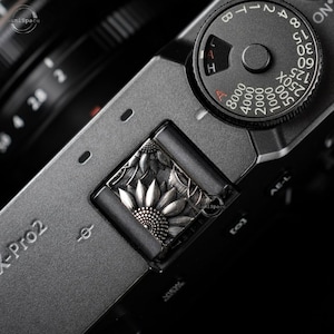 May include: A close-up of a black camera with a silver decorative plate featuring a sunflower design. The camera has a dial with the numbers 125, 250X, 500, 1000, 2000, 4000, and 8000. The camera also has a dial with the letters A, H, and L.