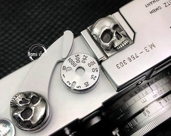 May include: A silver camera with a skull-shaped knob and a dial with numbers from 10 to 5000. The camera has the text "M3-756 303" and "TZ GMBH AR GERMANY" printed on it.