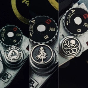 以下が含まれることがあります： 異なるデザインのカメラレンズキャップ3つ。最初のキャップは、「危険 高電圧」と書かれた白黒の警告標識。2つ目のキャップは、白黒の松の木の画像。3つ目のキャップは、銀と黒のタコと頭蓋骨の画像。