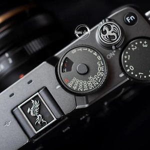 May include: A black camera with a silver dial that has the numbers 8, 1, 2, 4, 8, 15, 30, 60, 125, 250, 500, 1000, 2000, 4000, 8000 printed on it. The dial also has the letters A, B, T, and ON printed on it. The camera has a silver button with a phoenix design on it.