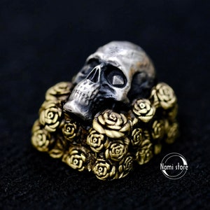 May include: A silver skull ring with a detailed design, resting on a bed of gold-toned roses. The skull has a dark, almost black, patina.