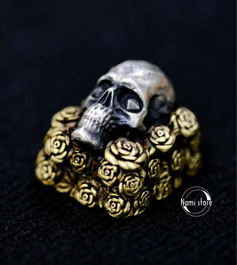 May include: A silver skull ring with a detailed design, resting on a bed of gold-toned roses. The skull has a dark, almost black, patina.