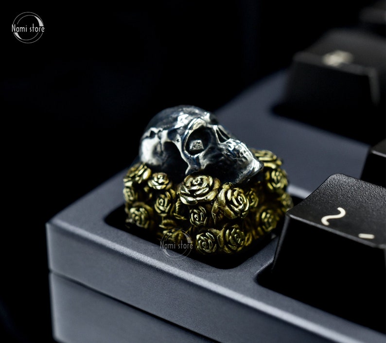 May include: A silver skull keycap with a gold rose design. The keycap is sitting on a black keyboard.