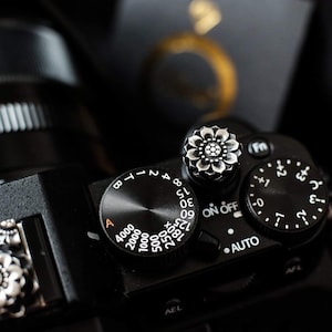 Puede incluir: Primer plano de una cámara negra con un dial plateado y una perilla con forma de flor. El dial tiene marcas para ajustes ISO, incluyendo 125, 180x, 4000, 2000, 1000, 500, 250, 15, 30 y 90/0. La perilla tiene un diseño de flor plateada con un centro negro.