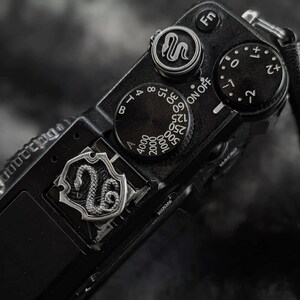 May include: Close-up of a black digital camera with silver accents. The camera features multiple dials and buttons, including a dial with numbers and a snake emblem. A leather strap is attached to the side. The camera is set against a blurred background.