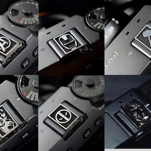 May include: Six different camera hot shoe covers with various superhero designs. The covers are made of metal and have a black finish. The designs include the Avengers logo, Iron Man, Thor's hammer, Deadpool, and Batman.