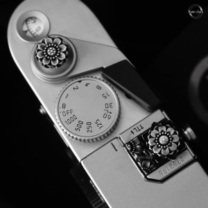 May include: A silver camera with a black dial and a silver knob with a floral design. The dial has numbers from 1 to 8 and the text "OFF", "1000", "500", "250", "125", "60", "30", "15", "8", "4", "2", and "1". The knob has a floral design with a silver center.
