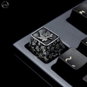 May include: A silver keycap with a black background and a maple leaf design. The keycap is surrounded by smaller maple leaf designs.
