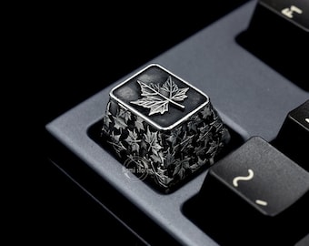 May include: A silver keycap with a black background and a maple leaf design. The keycap is surrounded by smaller maple leaf designs.