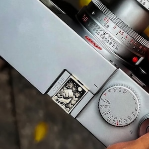 May include: A silver camera with a black and white engraved image of a forest scene on the top. The camera has a silver lens with a red ring and a silver dial with numbers and letters.