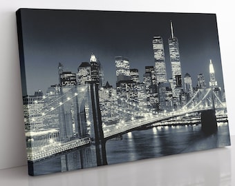 Op de afbeelding: Een zwart-witfoto van de Brooklyn Bridge 's nachts met de skyline van New York City op de achtergrond. De brug is verlicht met lampen, en de skyline van de stad is ook verlicht.