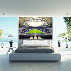 May include: A modern bedroom featuring a large canvas print of a football stadium. The stadium is empty and has a green pitch. The bedroom has a large bed with grey bedding and a white headboard. There are two lamps on the bedside tables and a window with a view of the sea.