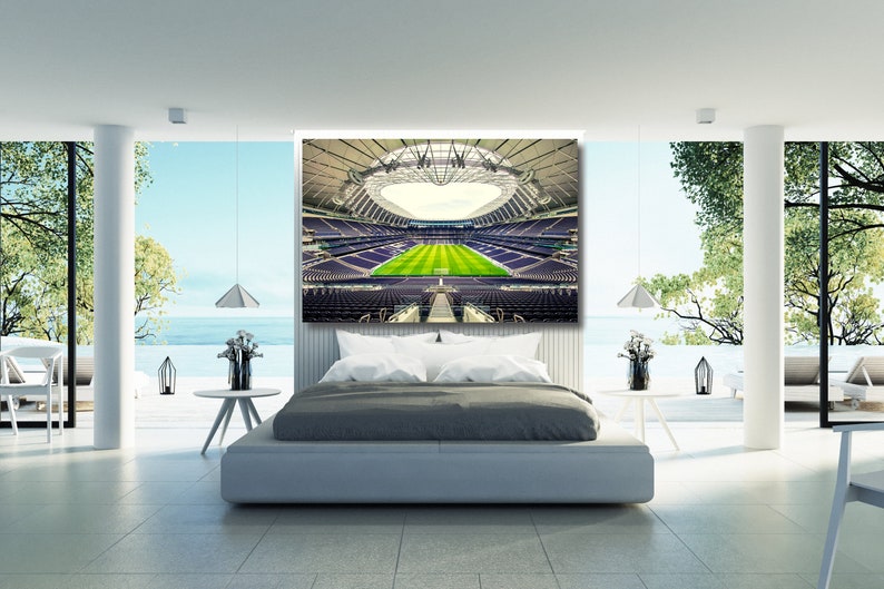 May include: A modern bedroom featuring a large canvas print of a football stadium. The stadium is empty and has a green pitch. The bedroom has a large bed with grey bedding and a white headboard. There are two lamps on the bedside tables and a window with a view of the sea.