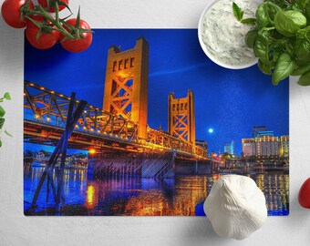 May include: A rectangular glass cutting board featuring a nighttime view of the Tower Bridge in Sacramento, California, illuminated with golden lights against a deep blue sky. The image is surrounded by fresh tomatoes, herbs, and a bowl of dip.