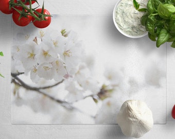 May include: A white glass cutting board with a floral design featuring white cherry blossoms. The board is surrounded by various ingredients, including tomatoes, garlic, basil, and a white sauce.