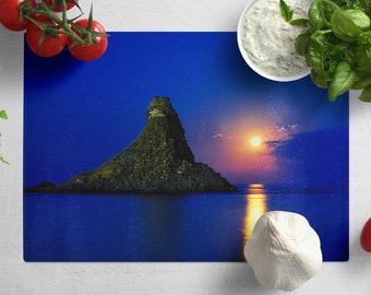 Peut inclure: Une planche à découper en verre bleu avec une image panoramique d'une île rocheuse dans l'océan au coucher du soleil. Le soleil se couche derrière l'île, projetant une lueur dorée sur l'eau. La planche à découper est entourée d'ingrédients frais, notamment des tomates, du basilic et de l'ail.