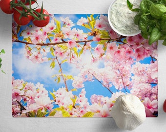 May include: A pink and white floral design kitchen cutting board with a blue sky background. The board is surrounded by various kitchen ingredients, including tomatoes, garlic, and herbs.