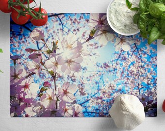 May include: A close-up of a cutting board with a floral design featuring pink and white cherry blossoms against a blue sky background. The cutting board is surrounded by various kitchen items, including a bowl of dip, tomatoes, and garlic.